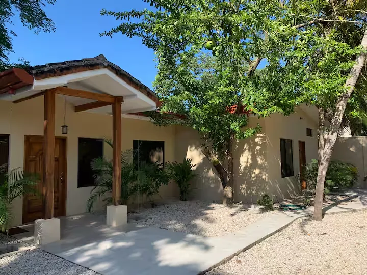 The two Casa Jícaro units, cream walls and wooden porch posts, under a jícaro tree in a gravel courtyard.